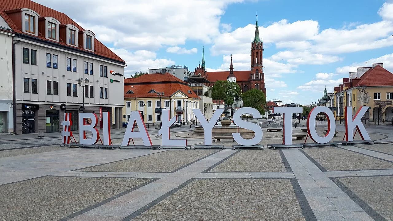 A wide view of the Bialystok letter sculpture with an old town square, buildings, and a church with a tall steeple in the background.