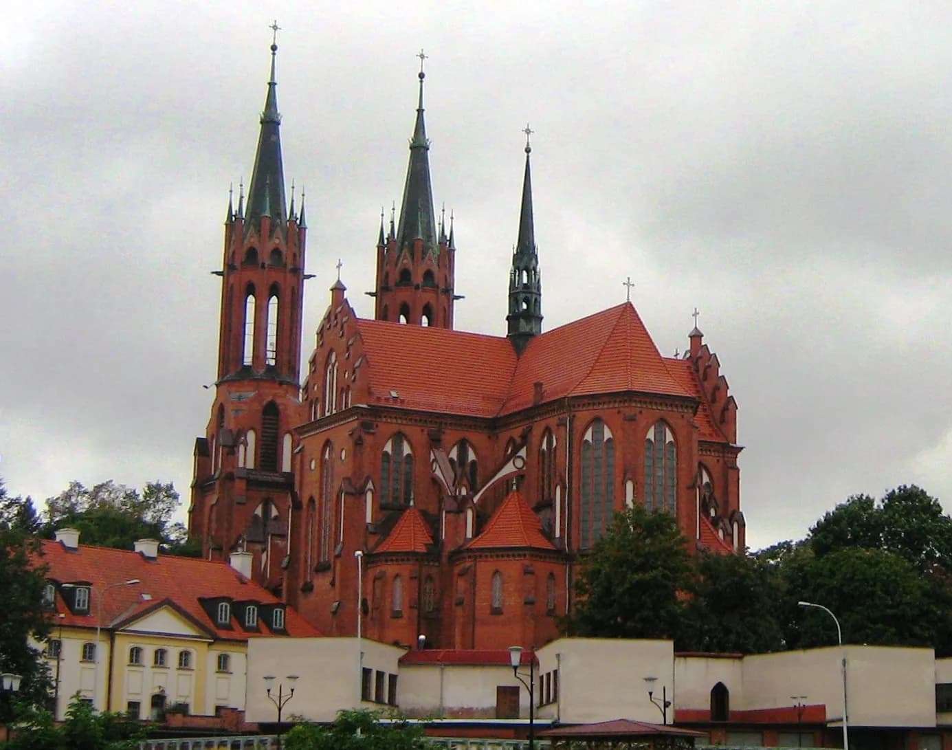 A large red brick cathedral with multiple spires stands against a cloudy sky, with smaller buildings and trees visible at its base.